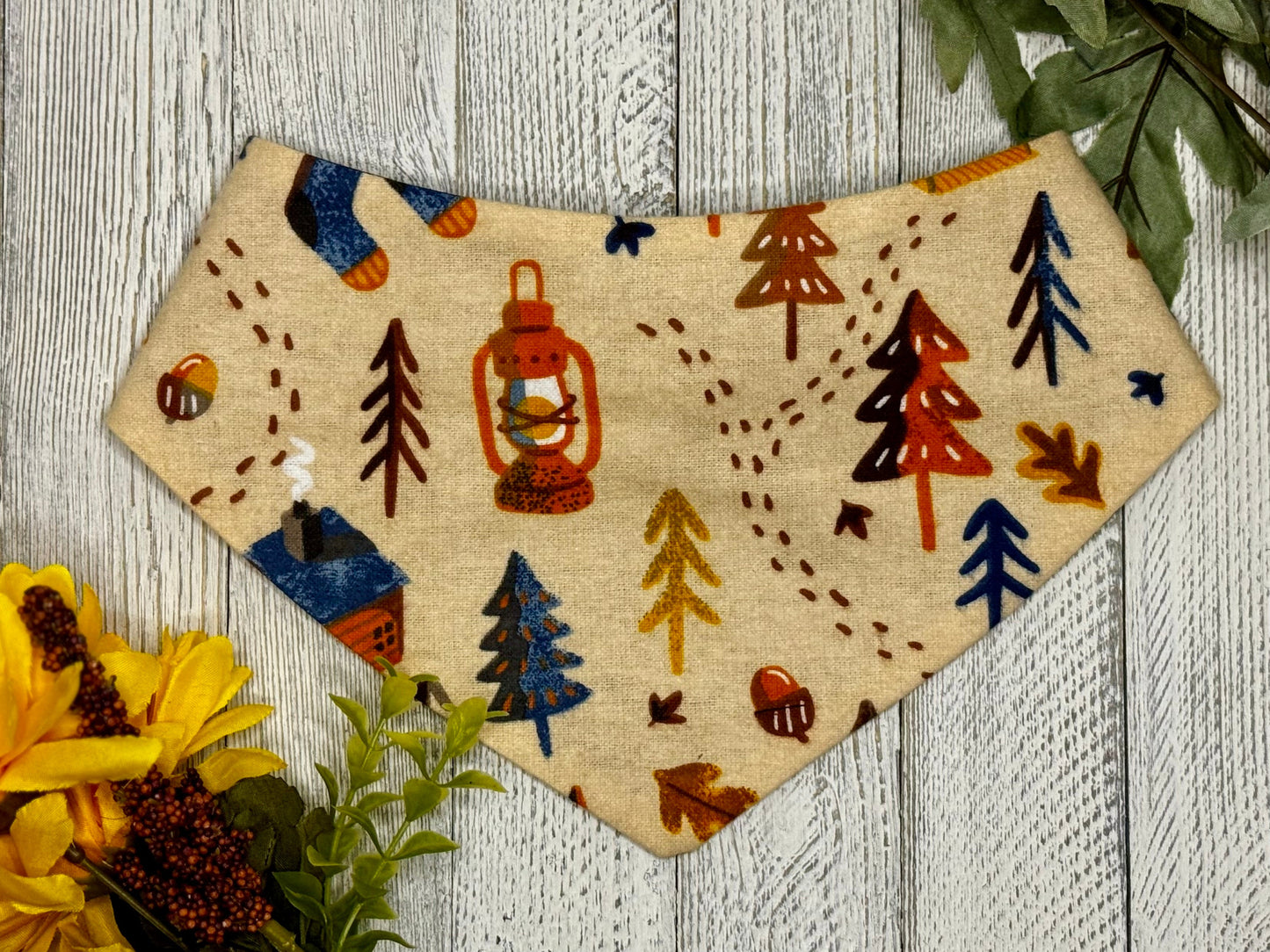 A pet bandana with a tan background featuring a camping pattern with trees, leaves, cabins, acorns, and lanterns, displayed on a wooden surface with flowers for decoration. 