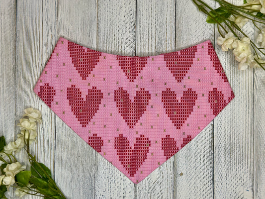 A pet bandana with a pink background featuring a heart cross stitch pattern, displayed on a wooden surface with flowers for decoration. 