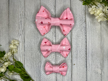 Pet bow ties (3 different sizes) with a pink background featuring a red and white arrow pattern, displayed on a wooden surface with flowers for decoration. 