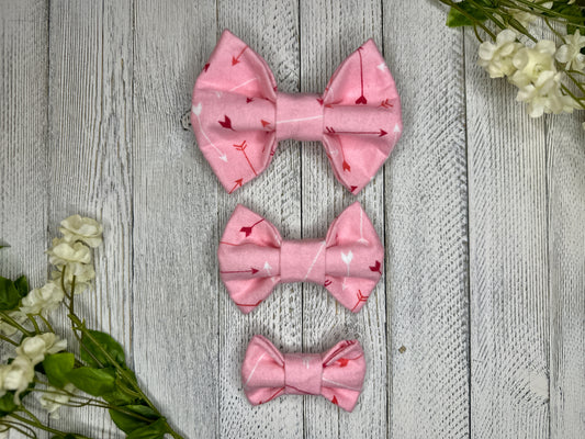 Pet bow ties (3 different sizes) with a pink background featuring a red and white arrow pattern, displayed on a wooden surface with flowers for decoration. 