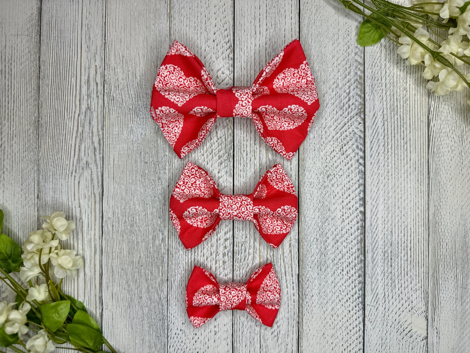 Three pet bow ties (small, medium, large) with a red background and white heart pattern displayed on a wooden surface with flowers for decoration.