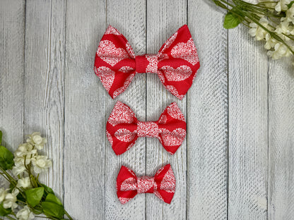 Three pet bow ties (small, medium, large) with a red background and white heart pattern displayed on a wooden surface with flowers for decoration.