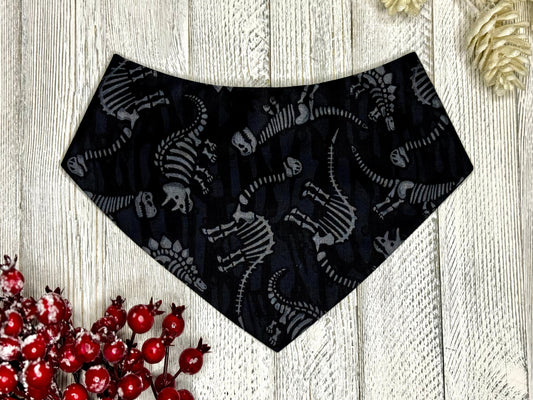 A black pet bandana with dinosaur fossils pattern, displayed on a wooden surface.