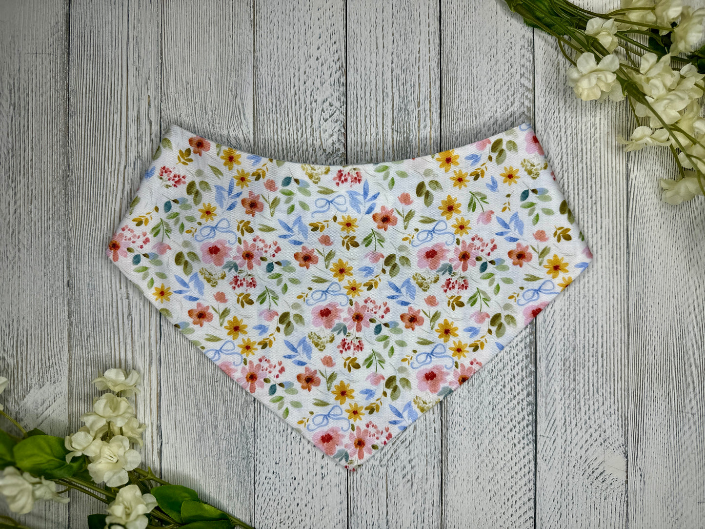 A pet bandana with a white background featuring a flower pattern with blue bows, displayed on a wooden surface with flowers for decoration.