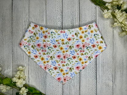 A pet bandana with a white background featuring a flower pattern with blue bows, displayed on a wooden surface with flowers for decoration.