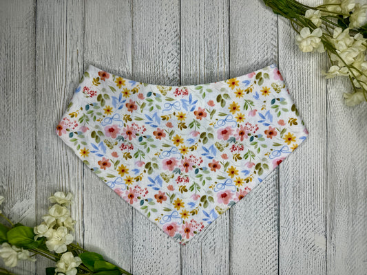 A pet bandana with a white background featuring a flower pattern with blue bows, displayed on a wooden surface with flowers for decoration.