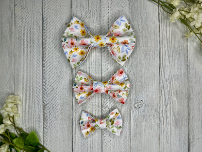 Three pet bow ties (small, medium, large) with a white background and wildflower pattern displayed on a wooden surface with flowers for decoration. 