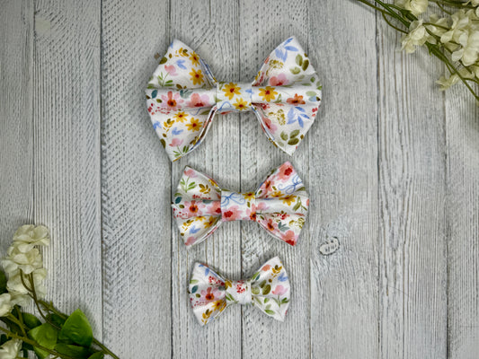 Three pet bow ties (small, medium, large) with a white background and wildflower pattern displayed on a wooden surface with flowers for decoration. 