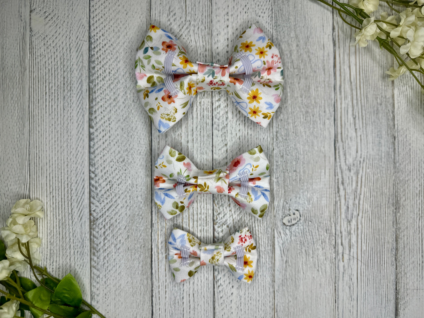 Picture of the backside of pet bow ties showing the elastic straps that fit around a collar. Displayed on a wooden surface with flowers for decoration. 