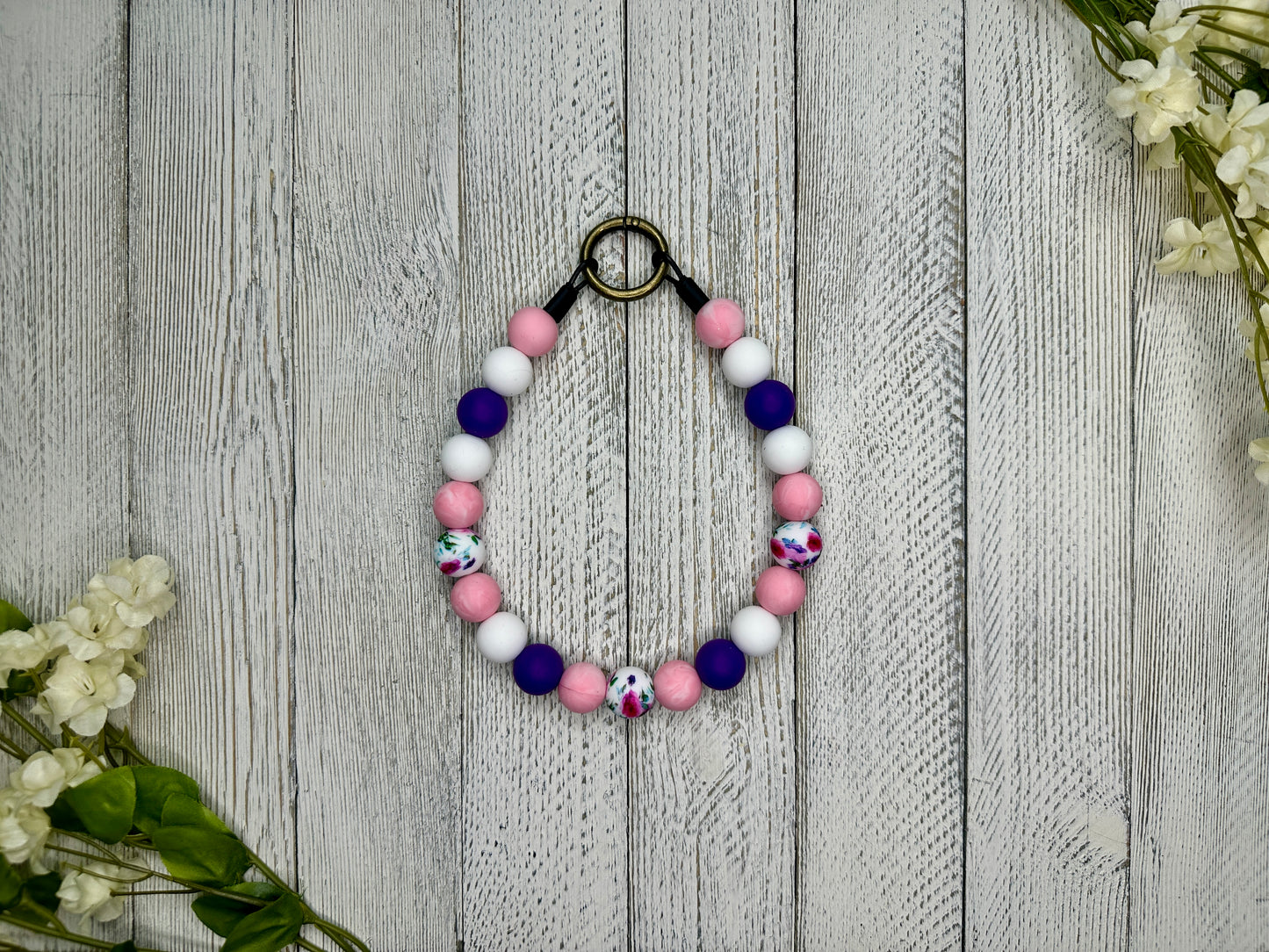 Beaded pet necklace with a purple, pink, and white pattern including flowers. Displayed on a wooden surface with flowers for decoration.