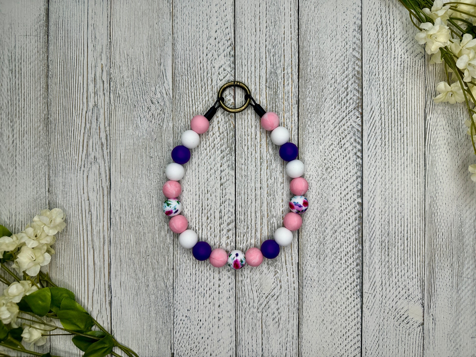 Beaded pet necklace with a purple, pink, and white pattern including flowers. Displayed on a wooden surface with flowers for decoration.