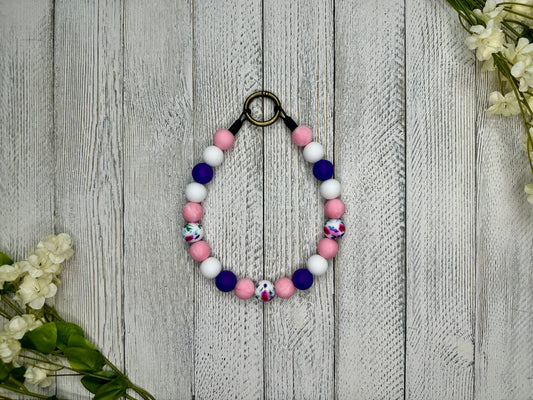 Beaded pet necklace with a purple, pink, and white pattern including flowers. Displayed on a wooden surface with flowers for decoration.