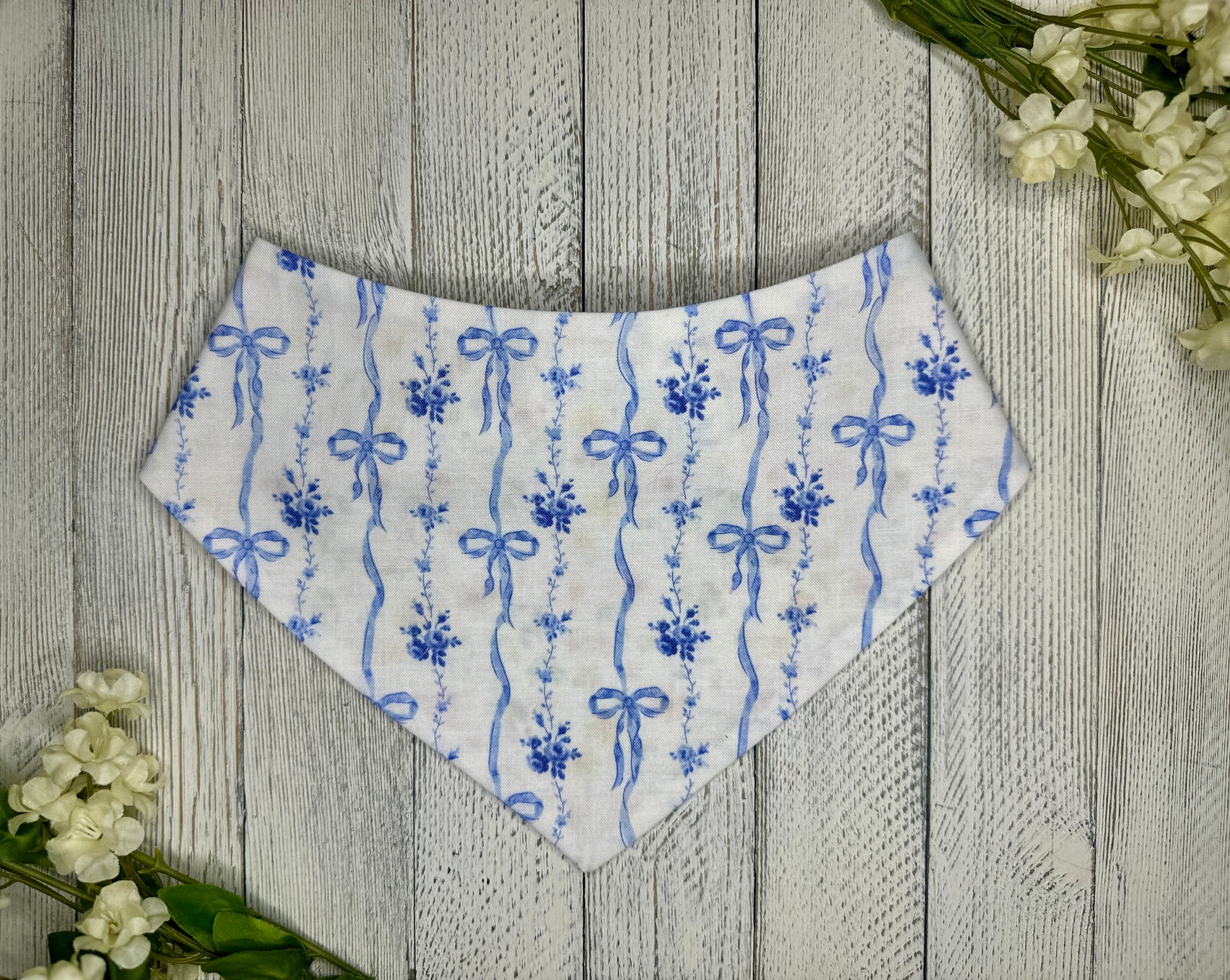 A pet bandana with a white background featuring dainty blue bows, displayed on a wooden surface with flowers for decoration. 
