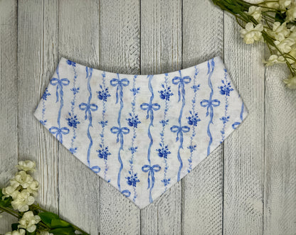A pet bandana with a white background featuring dainty blue bows, displayed on a wooden surface with flowers for decoration. 