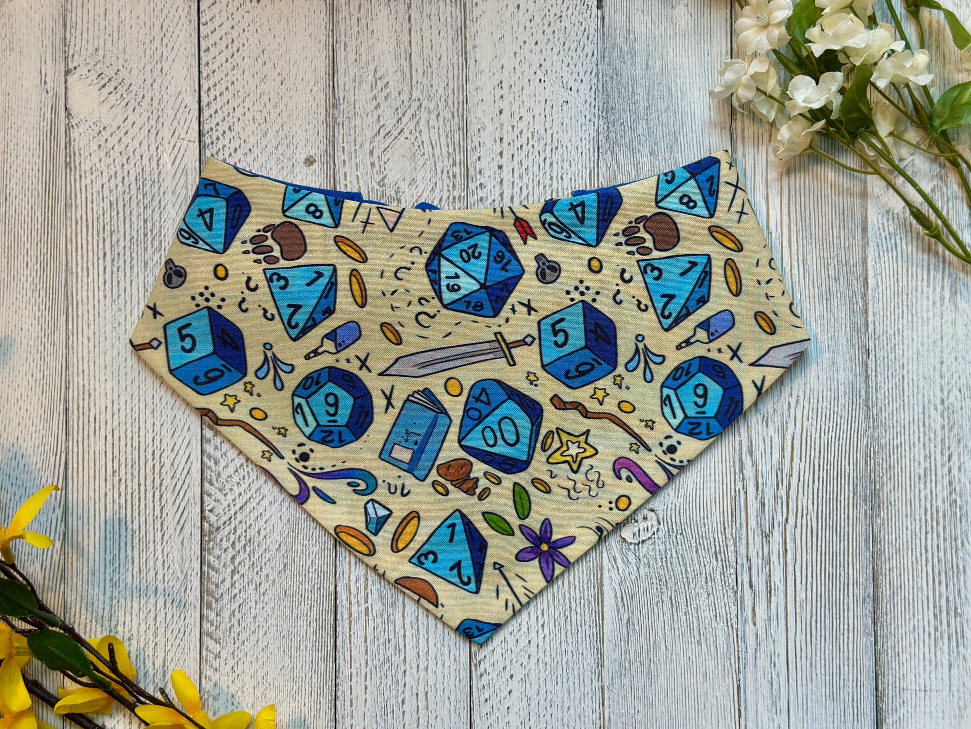 Pet bandana with blue dice and gaming-themed design on a wooden surface with flowers.