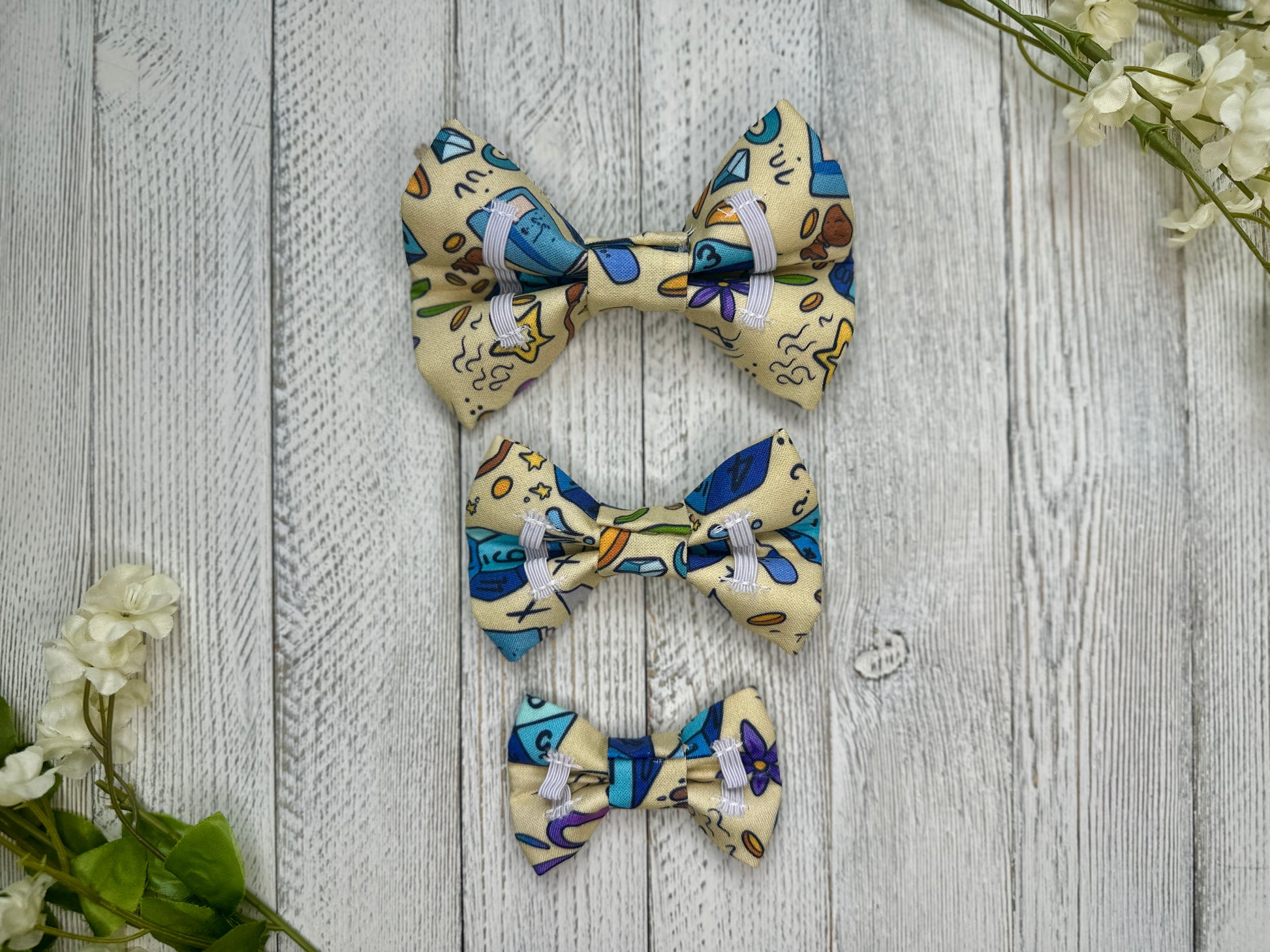 Picture of the backside of pet bow ties showing the elastic straps that fit around a collar. Displayed on a wooden surface with flowers for decoration. 
