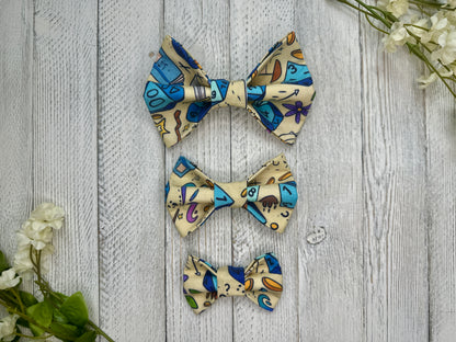 Three pet bow ties (small, medium, large) with with a dice and dungeon pattern, displayed on a wooden surface with flowers for decoration. 