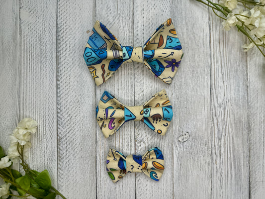 Three pet bow ties (small, medium, large) with with a dice and dungeon pattern, displayed on a wooden surface with flowers for decoration. 