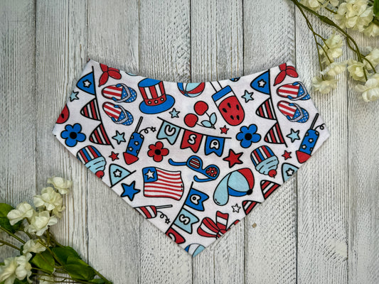 A pet bandana with a white background featuring red and blue pattern with a USA theme, displayed on a wooden surface with flowers for decoration. 