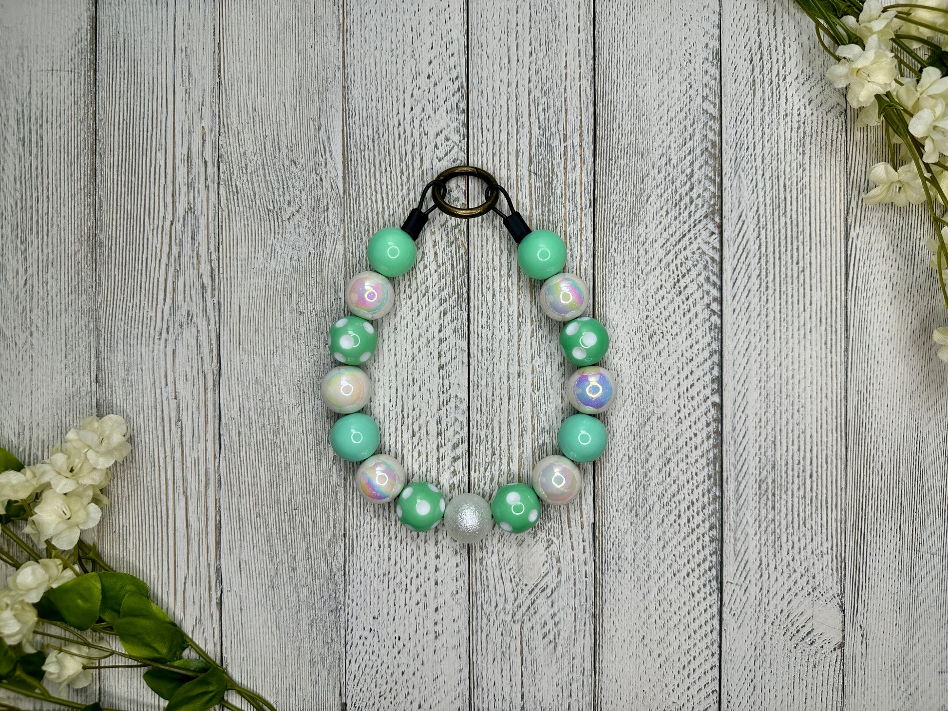 Beaded pet necklace with a mint and white pattern including polka dots. Displayed on a wooden surface with flowers for decoration. 