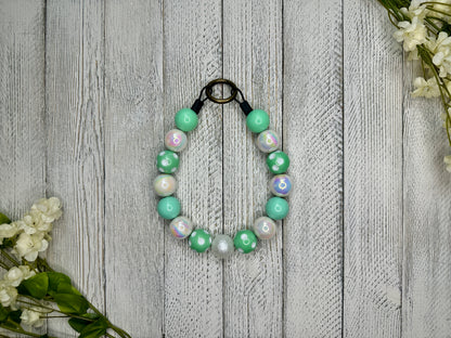 Beaded pet necklace with a mint and white pattern including polka dots. Displayed on a wooden surface with flowers for decoration. 