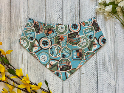 A pet bandana with a light blue background featuring patches of various pet related items, displayed on a wooden surface with flowers for decoration. 
