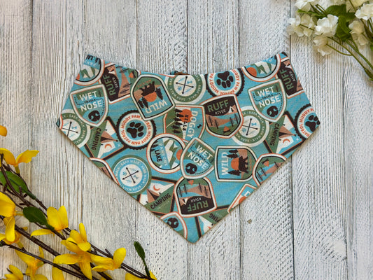 A pet bandana with a light blue background featuring patches of various pet related items, displayed on a wooden surface with flowers for decoration. 