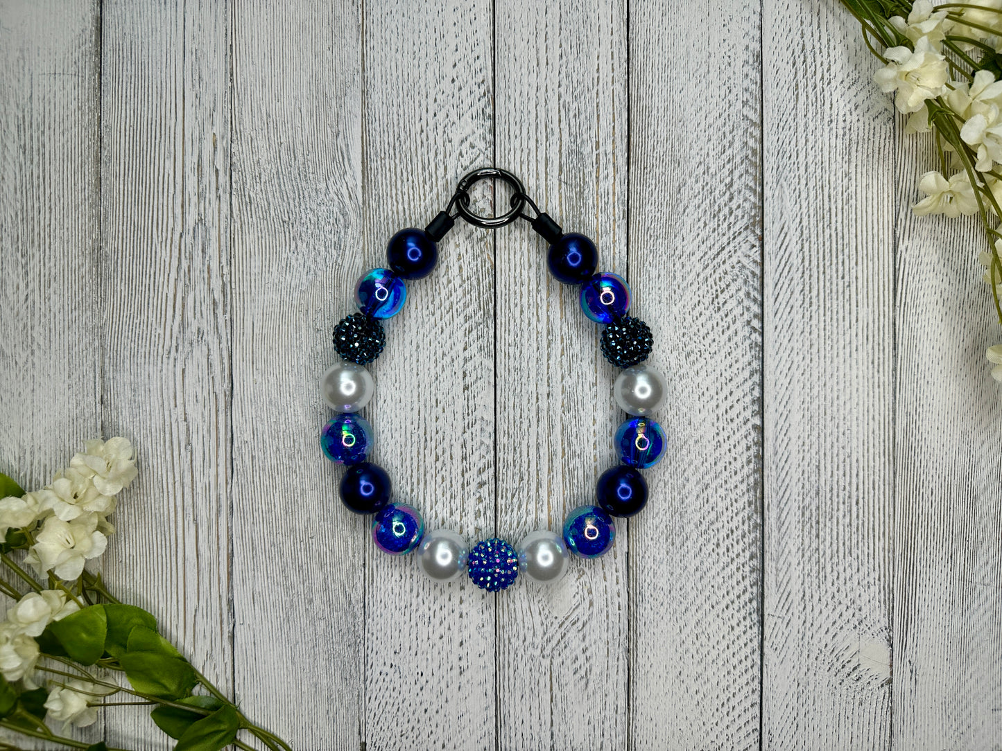 Beaded pet necklace with a navy blue pattern including sparkles. Displayed on a wooden surface with flowers for decoration.