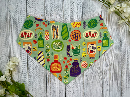 A pet bandana with a green background featuring a grillin' and BBQ theme, displayed on a wooden surface with flowers for decoration.