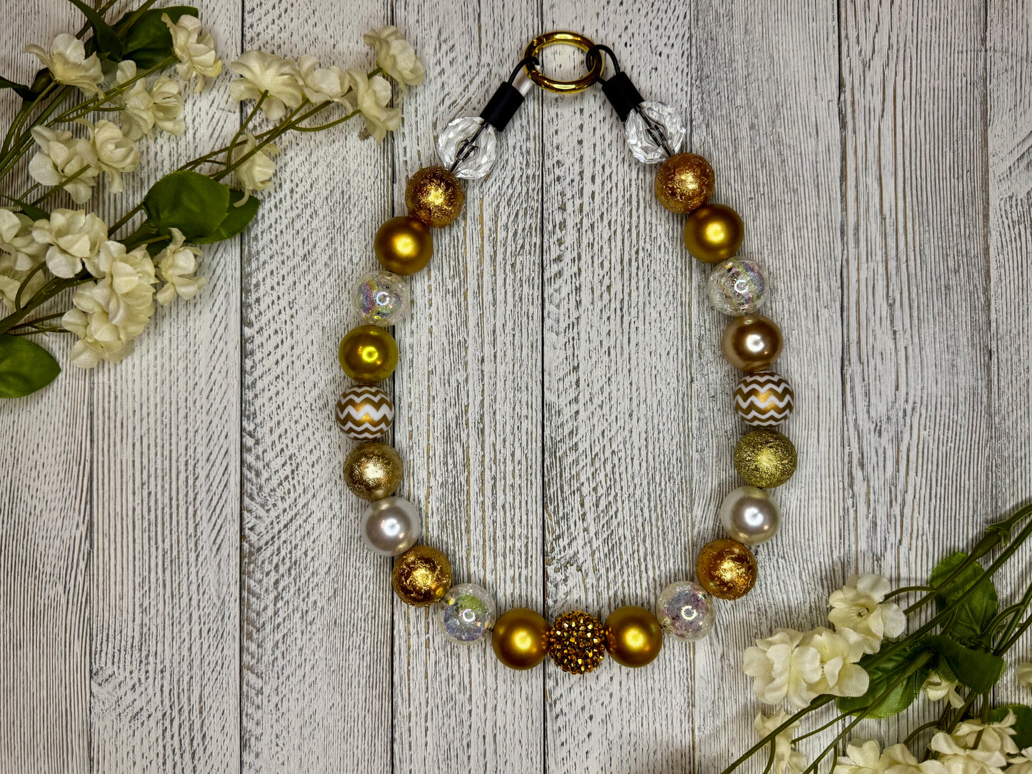 Beaded dog necklace with a gold pattern including zig zags. Displayed on a wooden surface with flowers for decoration.