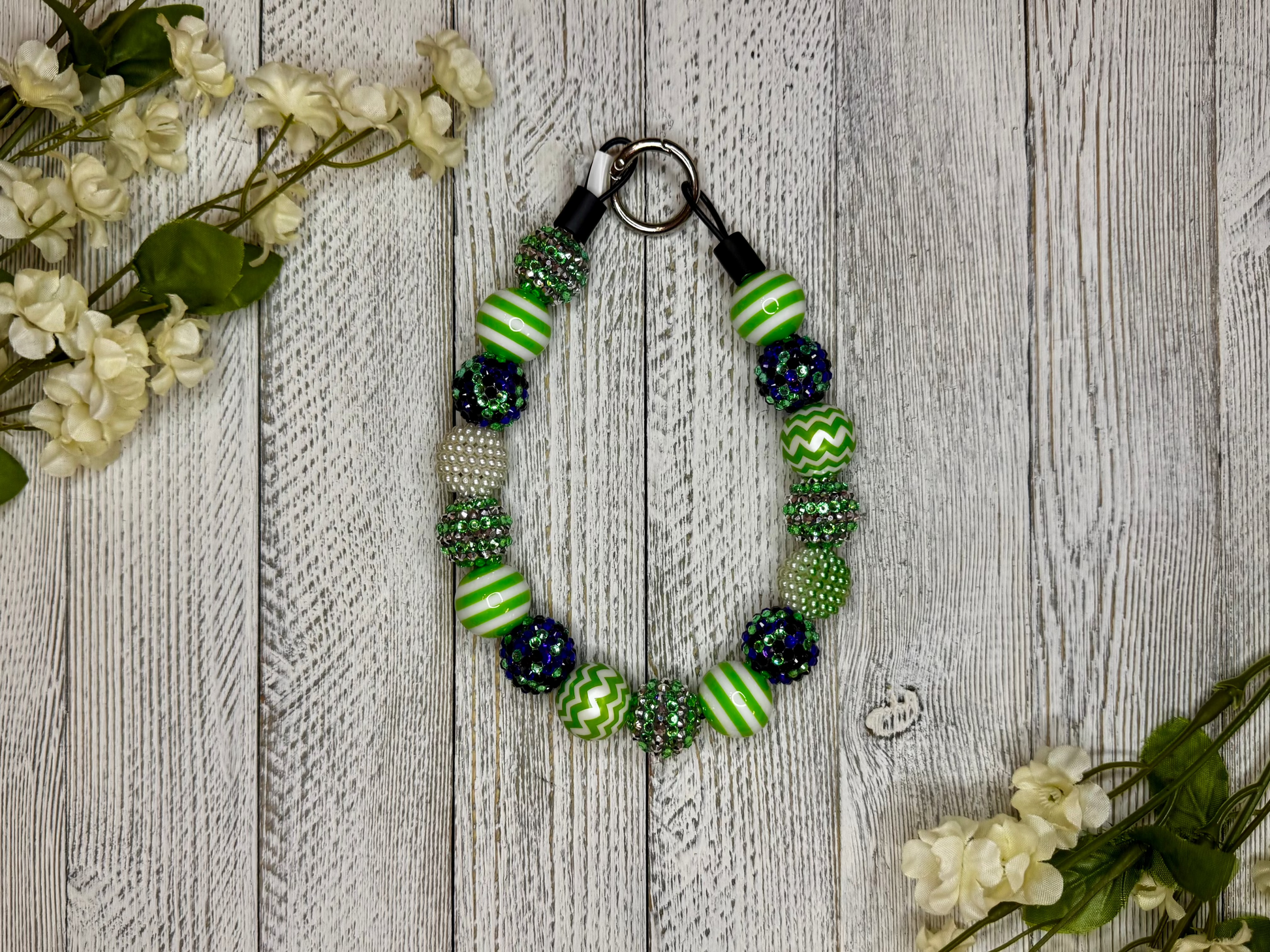 Beaded dog necklace with a green and purple pattern including stripes. Displayed on a wooden surface with flowers for decoration.