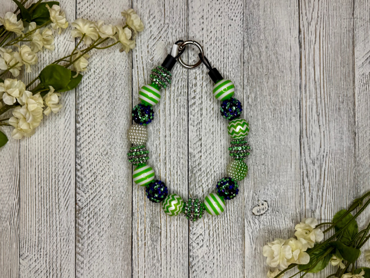 Beaded dog necklace with a green and purple pattern including stripes. Displayed on a wooden surface with flowers for decoration.