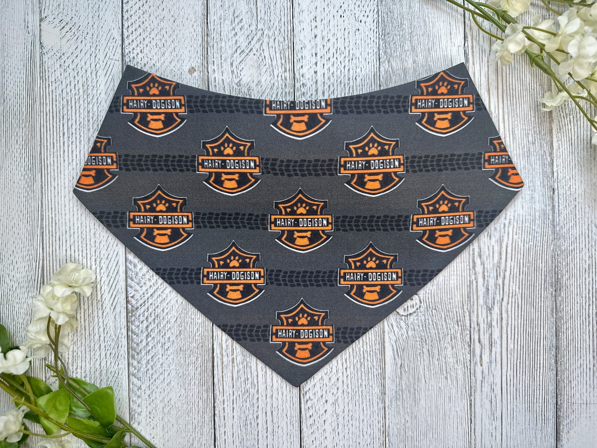 A pet bandana with a black background featuring a tire track and motorcycle logo, displayed on a wooden surface with flowers for decoration. 