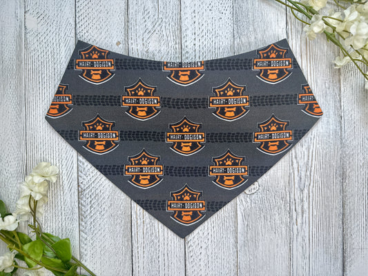 A pet bandana with a black background featuring a tire track and motorcycle logo, displayed on a wooden surface with flowers for decoration. 