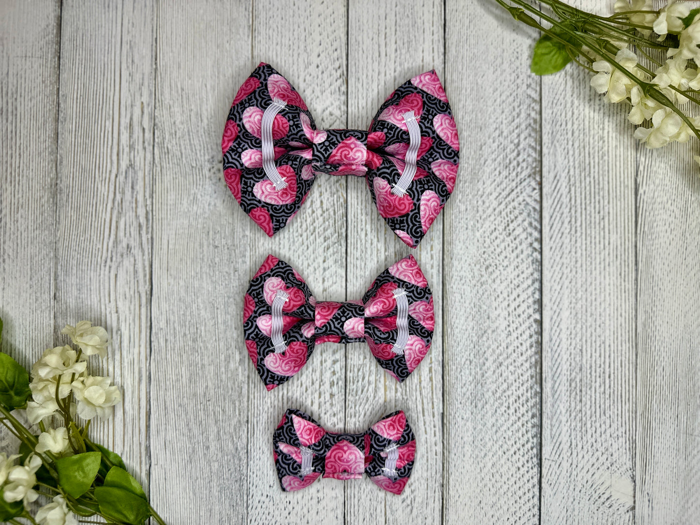Picture of the backside of pet bow ties showing the elastic straps that fit around a collar. Displayed on a wooden surface with flowers for decoration. 