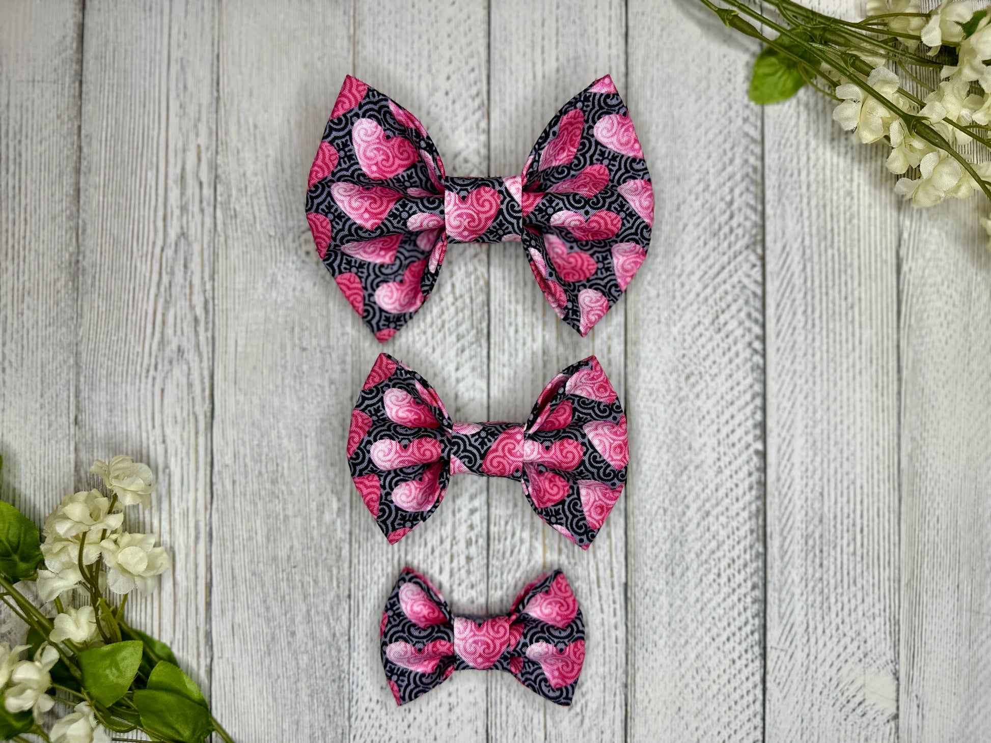 Three pet bow ties (small, medium, large) with a black and grey background and pink hearts displayed on a wooden surface with flowers for decoration. 