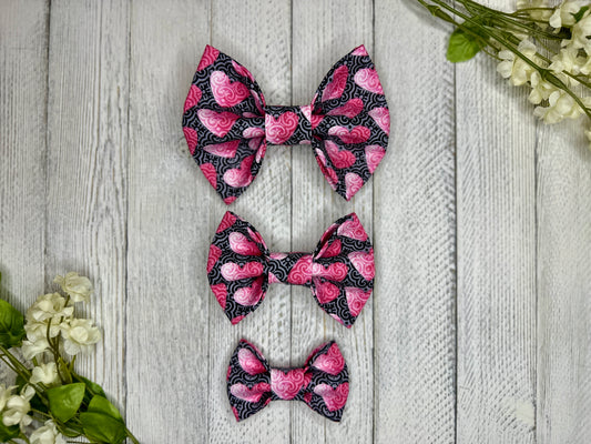 Three pet bow ties (small, medium, large) with a black and grey background and pink hearts displayed on a wooden surface with flowers for decoration. 