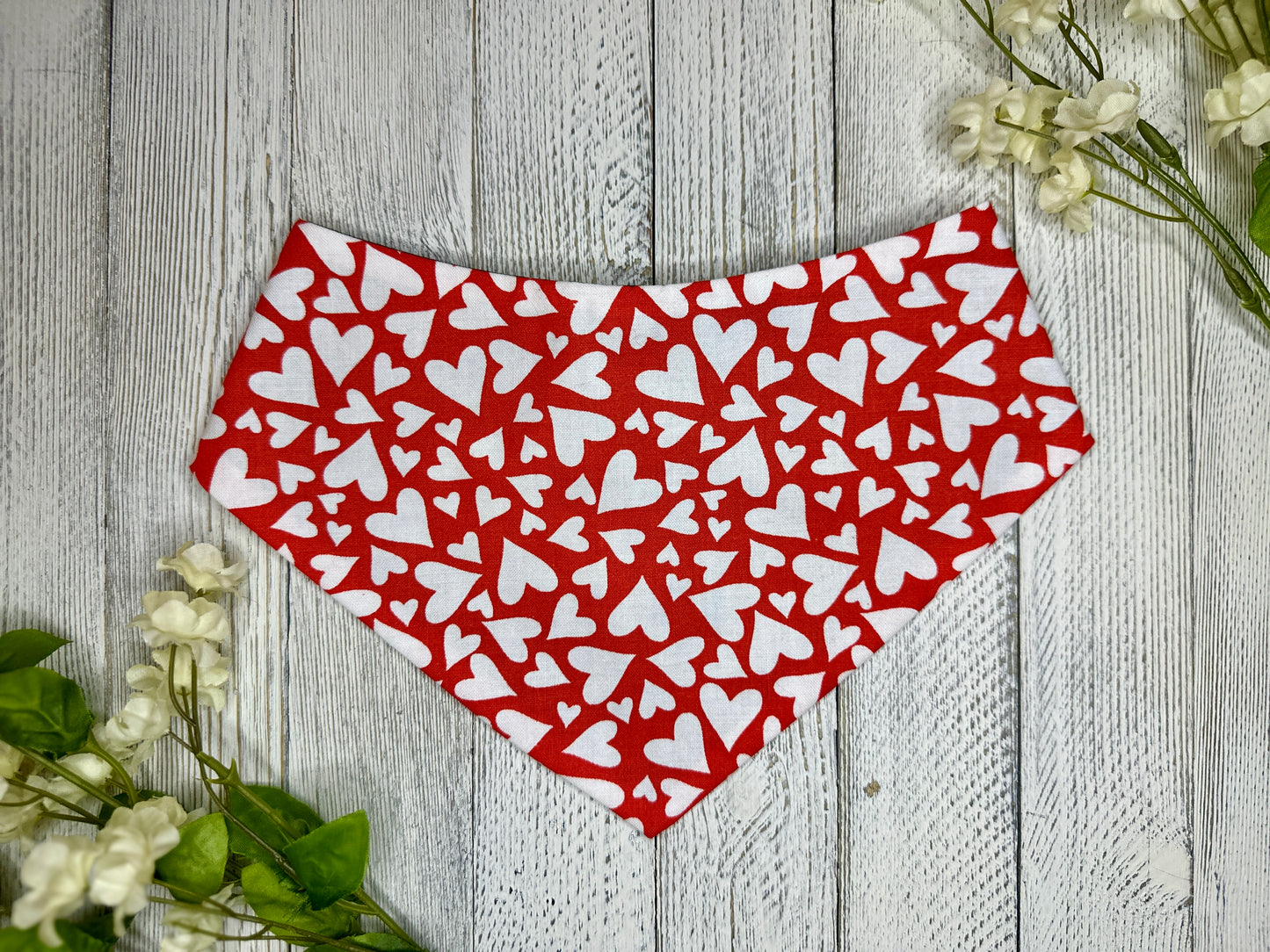 A pet bandana with a red background featuring a white heart pattern, displayed on a wooden surface with flowers for decoration. 