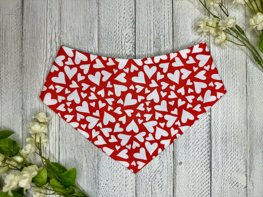 A pet bandana with a red background featuring a white heart pattern, displayed on a wooden surface with flowers for decoration. 