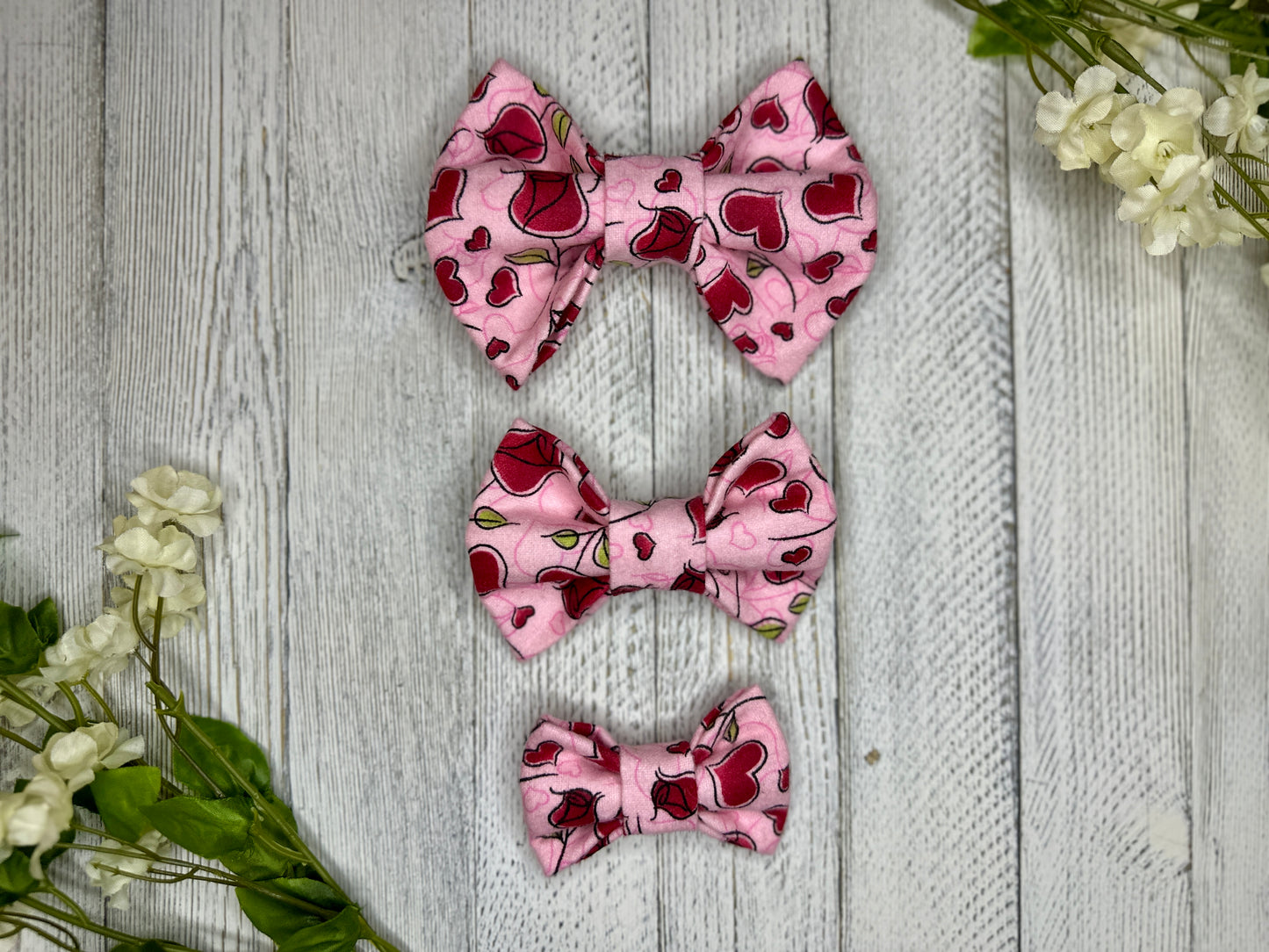 A pet bow tie with a pink background featuring a rose pattern with red hearts, displayed on a wooden surface with flowers for decoration. 