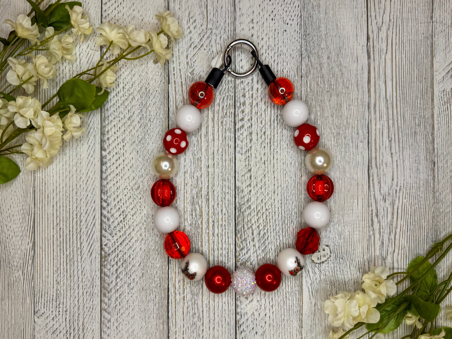 Beaded dog necklace with a red and white pattern including reindeer. Displayed on a wooden surface with flowers for decoration.