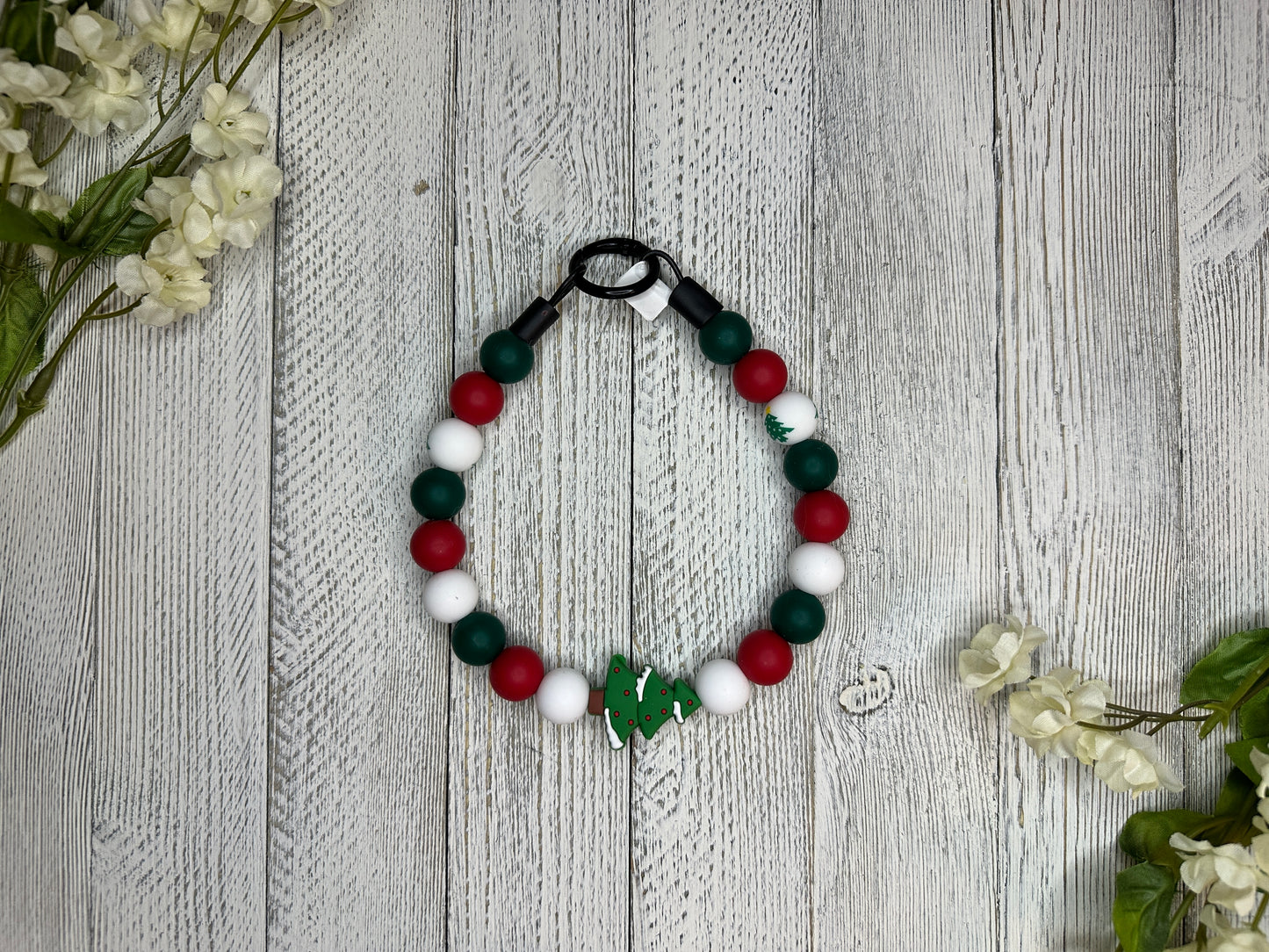 Beaded dog necklace with a red, green, and white pattern including Christmas trees. Displayed on a wooden surface with flowers for decoration.