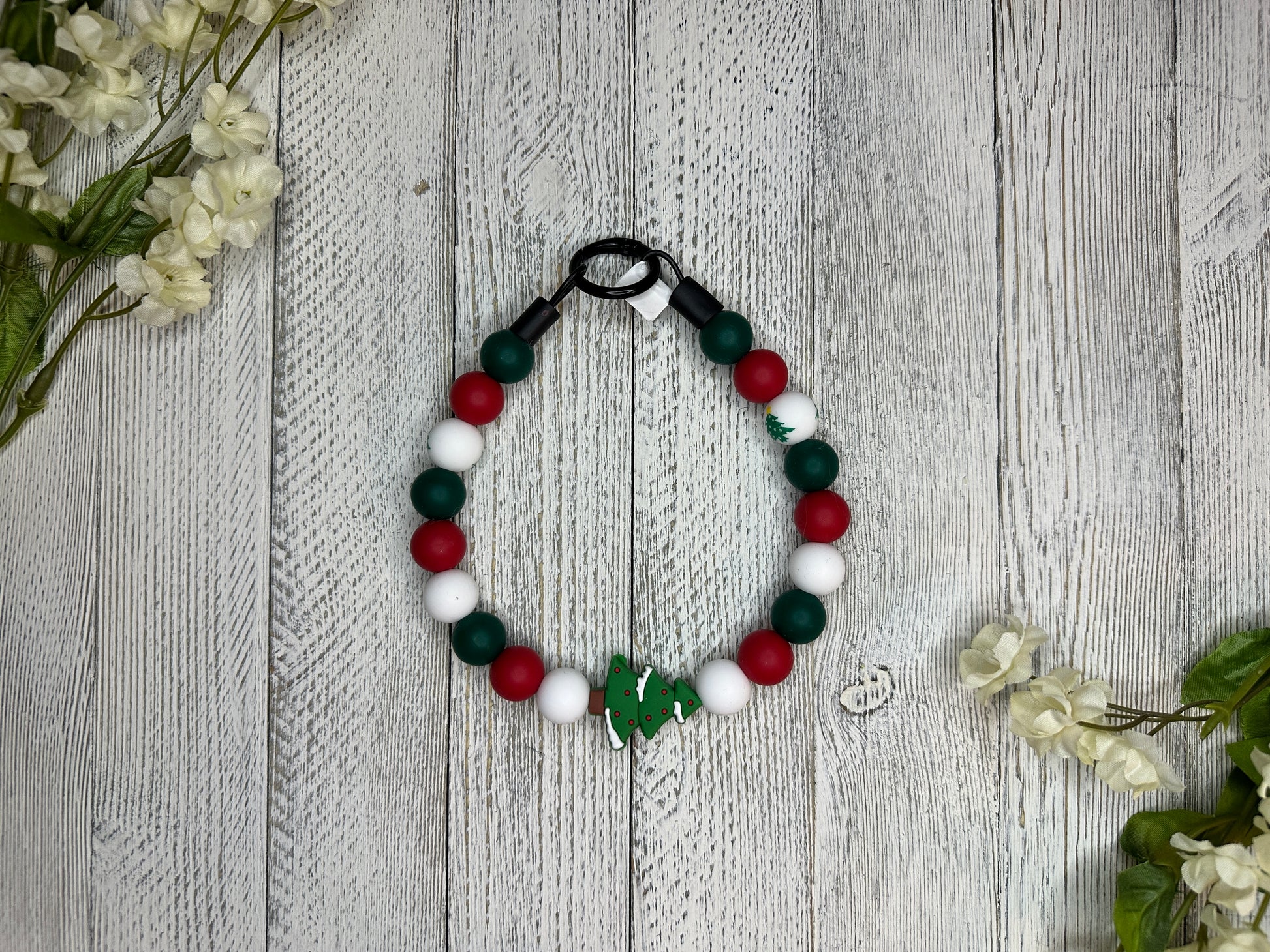 Beaded dog necklace with a red, green, and white pattern including Christmas trees. Displayed on a wooden surface with flowers for decoration.