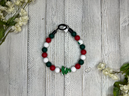 Beaded dog necklace with a red, green, and white pattern including Christmas trees. Displayed on a wooden surface with flowers for decoration.