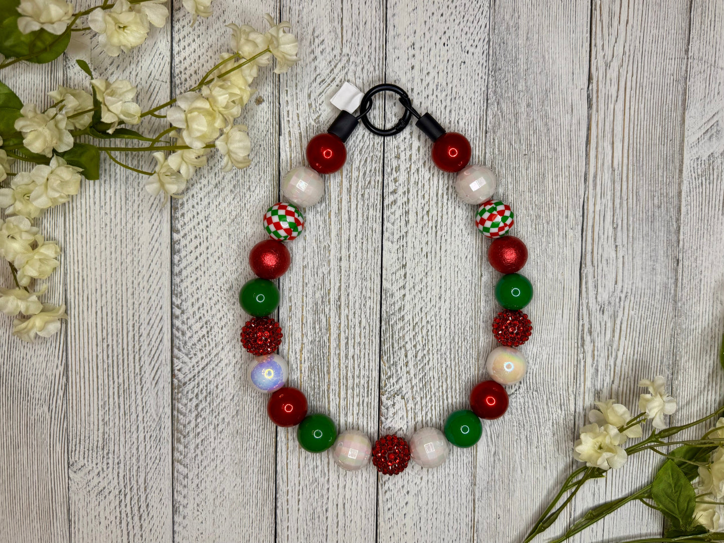 Beaded dog necklace with a red, green, and white pattern including checkers. Displayed on a wooden surface with flowers for decoration.