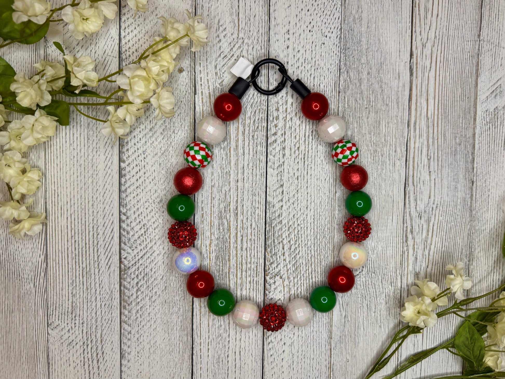 Beaded dog necklace with a red, green, and white pattern including checkers. Displayed on a wooden surface with flowers for decoration.