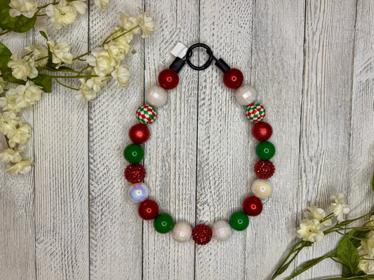 Beaded dog necklace with a red, green, and white pattern including checkers. Displayed on a wooden surface with flowers for decoration.