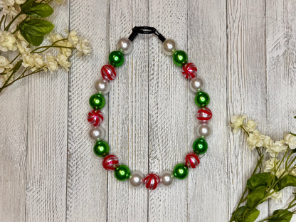 Beaded dog necklace with a red, green, and white pattern including candy cane stripes. Displayed on a wooden surface with flowers for decoration.