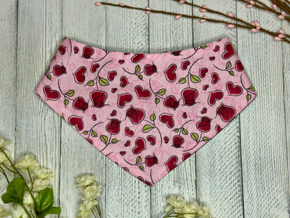 A pet bandana with a pink background featuring a rose pattern with red hearts, displayed on a wooden surface with flowers for decoration. 