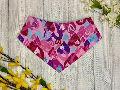 A pet bandana with a pink background featuring varying colors of hearts (pink, red, white, purple, blue) displayed on a wooden surface with flowers for decoration. 